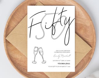 May include: A white invitation card with the word "Fifty" in a stylish script, celebrating a birthday. The card includes details like the date, time, and location, with two champagne glasses illustrated at the bottom. The card is placed on a wooden tray.