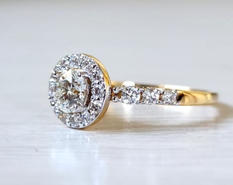 Op de afbeelding: Een gouden verlovingsring met een ronde centrale diamant omgeven door een halo van kleinere diamanten. De band is ook bezet met kleinere diamanten.