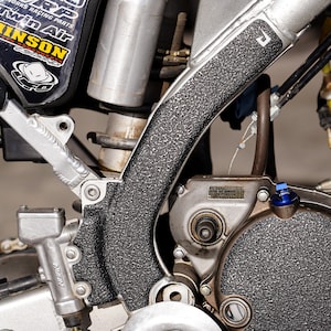 May include: Close-up of a motorcycle frame with a black textured protective layer. The frame is made of silver metal and has a bolt in the lower right corner. The protective layer is covering the frame and has a rough texture.