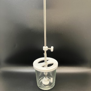May include: A clear glass jar with a white plastic lid and a central mechanism. A long, thin metal rod extends from the lid, connected to a white knob at the top. The jar has a white plastic base and a spring mechanism.