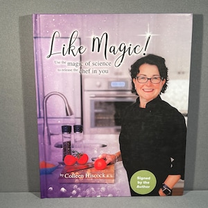 Könnte beinhalten: Ein lila-weißes Kochbuch mit dem Titel "Like Magic!" und dem Text "Use the magic of science to release the chef in you" von Colleen Hiscock. Eine Frau in einem schwarzen Shirt hält eine rote Tomate in der Hand. Das Buch ist vom Autor signiert.