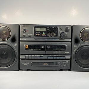 May include: A vintage Panasonic stereo system in dark grey, featuring two speakers with gold-toned accents. It includes a CD player, dual cassette decks, and an AM/FM radio. The display shows "TAPE" and "40 POS".