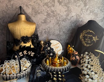 Puede incluir: Una colección de accesorios de boda en negro y dorado que incluye un maniquí drapeado en tul negro, una cesta con detalles de perlas, una bandeja dorada con un adorno central, una tela negra con bordados dorados y tocados florales.