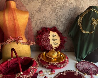 Puede incluir: Una colección de accesorios de boda en color burdeos y dorado que incluye un maniquí con velo, una cesta de terciopelo, un cartel circular con el texto Ayse Gelinin Kinasi, una bandeja dorada con velas, un abanico de encaje y piezas de tela.