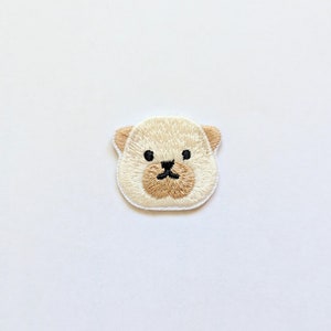 Può includere: Toppa ricamata raffigurante la faccia di un cucciolo di orso beige chiaro, con occhi e naso neri. Le orecchie e il muso dell'orso sono di un beige più scuro. La toppa è progettata per essere stirata o cucita sul tessuto.