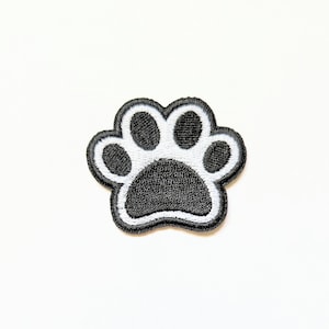 Puede incluir: Parche bordado con estampado de huella de pata en blanco y negro. El parche presenta un diseño detallado de una pata con cinco dedos, delineados con costuras blancas. La pata es un símbolo de mascotas y animales.