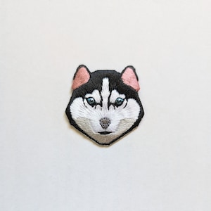 May include: Embroidered patch of a husky dog's face with black fur, white markings, and blue eyes. The dog has a pink nose and pink ears.