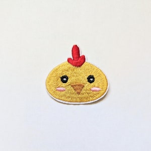 Pode incluir: Um pintinho amarelo bordado com uma crista vermelha e olhos pretos. O pintinho tem um bico pequeno e bochechas rosadas.