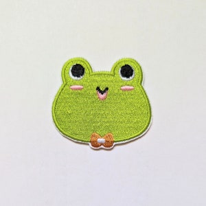 Puede incluir: Parche de rana verde bordado con ojos negros, mejillas rosadas y una pequeña corbata de lazo naranja.