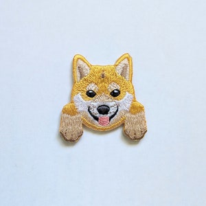 Può includere: Toppa ricamata raffigurante un cane Shiba Inu sorridente. La toppa ha una combinazione di colori giallo e bianco con occhi neri e una lingua rosa. Le zampe del cane sono estese, come se stesse sbirciando sopra qualcosa.