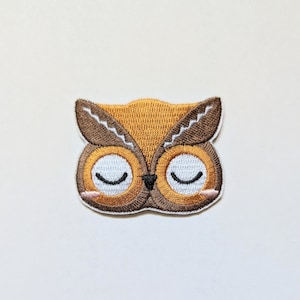 Op de afbeelding: Een geborduurde uil-patch met gesloten ogen. De uil is bruin en oranje en kan op stof worden genaaid.