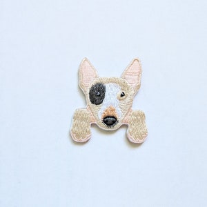 May include: Embroidered iron-on patch of a white and tan bull terrier dog with black eyes and a pink nose. The dog is looking directly at the viewer with its mouth closed.