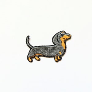 Pode incluir: Etiqueta bordada de um dachshund preto e castanho. O dachshund está virado para a direita e tem um corpo comprido e pernas curtas.