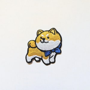 Könnte beinhalten: Ein gestickter Bügelaufkleber mit einem Cartoon-Shiba-Inu-Hund, der eine blaue Schleife trägt. Der Hund ist gelb mit weißen Markierungen und hat einen fröhlichen Ausdruck.