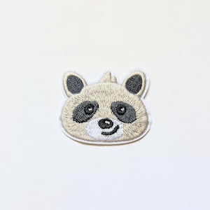 Puede incluir: Un parche blanco bordado con la cara de un mapache con ojos negros y nariz negra.