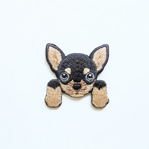 Op de afbeelding: Gestikt patch van een zwarte en bruine chihuahua met grote ogen, die omhoog kijkt en achter een muur vandaan kijkt.