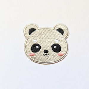 Puede incluir: Un parche de planchado bordado de color beige con un oso de dibujos animados con ojos negros y mejillas rosadas.