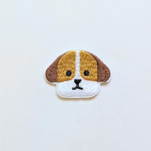 May include: Embroidered iron-on patch of a brown and white dog with black eyes. The dog has a simple, cartoon-like design.