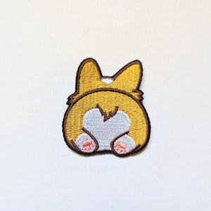 Peut inclure: Patch brodé représentant l'arrière-train d'un corgi de dessin animé. Le corgi est jaune avec des oreilles brunes et une queue blanche. Les pattes du corgi sont roses.
