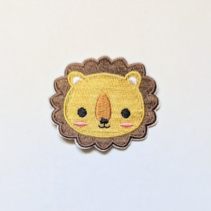 May include: A brown and yellow embroidered lion patch with a cute cartoon face. The lion has a brown mane, a yellow face, and black eyes.