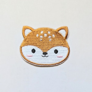 Puede incluir: Un parche bordado marrón con una cara de zorro blanca. El zorro tiene ojos negros, una pequeña nariz negra y mejillas rosadas.