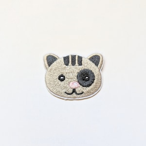 May include: An iron-on patch featuring an embroidered beige cat with black stripes and a black eye. The cat has a pink nose and a white smile.