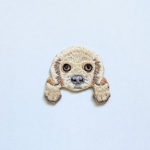 May include: A beige embroidered patch of a dog peeking over the top of a surface. The dog has brown eyes and a brown nose.