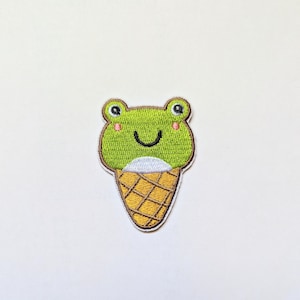 以下が含まれることがあります： 笑顔でピンクの頬をした緑色のカエルのアイスクリームコーンの刺繍パッチ