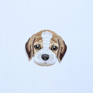 Może przedstawiać: Haftowana naszywka przedstawiająca pysk psa rasy beagle. Naszywka ma biało-brązową kolorystykę, ze szczegółowymi przeszyciami definiującymi cechy psa, w tym oczy, nos i uszy. Naszywka jest przeznaczona do naprasowywania lub przyszywania do tkaniny.