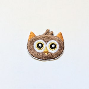Könnte beinhalten: Gestickter brauner Eulen-Patch mit weißen Augen und einem gelben Schnabel. Die Eule hat orangefarbene Ohren.