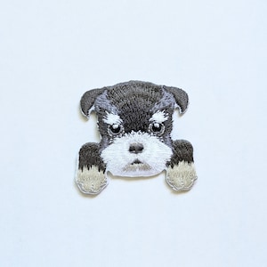 May include: Embroidered patch of a black and white miniature schnauzer puppy with brown ears and paws. The puppy is looking directly at the viewer with a serious expression.