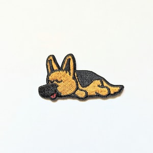 May include: Embroidered patch of a brown and black German Shepherd dog sleeping. The dog is lying on its side with its head resting on its paws.