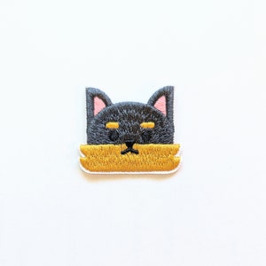 May include: Embroidered patch featuring a black cat face with pink inner ears and yellow accents. The cat's face is partially obscured by a yellow rectangular shape. The patch has a white border and is likely for clothing or accessories.