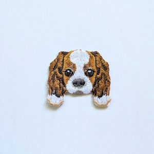 以下が含まれることがあります： キャバリア・キング・チャールズ・スパニエルの犬の刺繍パッチ。パッチは白と茶色の配色で、犬の特徴を生き生きとさせる詳細なステッチが施されています。生地にアイロンをかけたり、縫い付けたりするように設計されています。