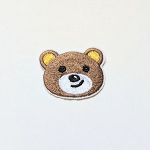 May include: Embroidered brown bear face patch with yellow ears. The bear has a white nose and a smiling mouth.