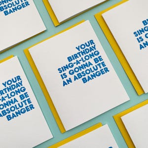 May include: A white card with a yellow border and blue text that reads "Your birthday sing-a-long is gonna be an absolute banger".