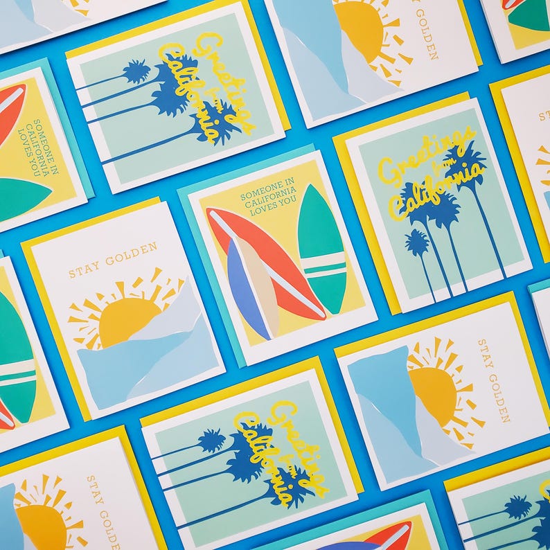 May include: A set of greeting cards with a California theme. The cards feature illustrations of palm trees, surfboards, and sunsets. The text on the cards includes "Greetings from California", "Stay Golden", and "Someone in California loves you".
