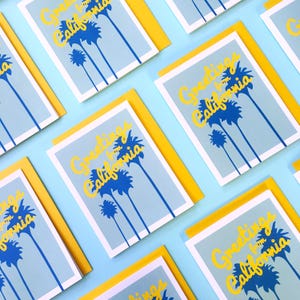 May include: A flat lay of greeting cards with a blue and yellow design. The cards feature the text "Greetings from California" and a graphic of palm trees. The cards are arranged on a light blue background.