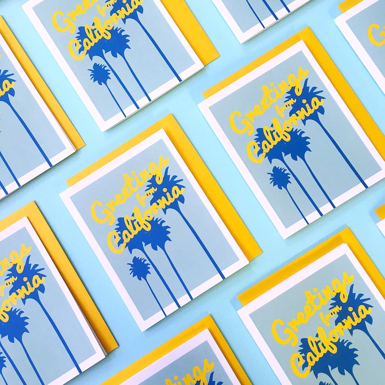 May include: A flat lay of greeting cards with a blue and yellow design. The cards feature the text "Greetings from California" and a graphic of palm trees. The cards are arranged on a light blue background.