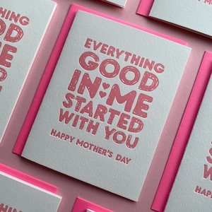 May include: Greeting cards with pink text on white paper, featuring the message "EVERYTHING GOOD IN ME STARTED WITH YOU" and "HAPPY MOTHER'S DAY". The cards have pink borders and are arranged on a pink surface.