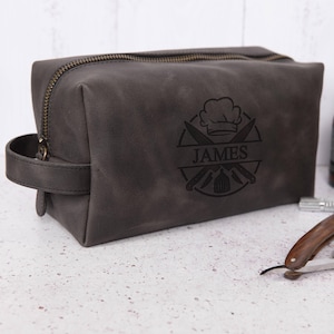 以下が含まれることがあります： ジッパー留めと革製のハンドルが付いたグレーの革製のトイレタリーバッグ。バッグには、シェフの帽子、交差したナイフ、そして「JAMES」という名前が刻まれたパーソナライズされたデザインが施されています。