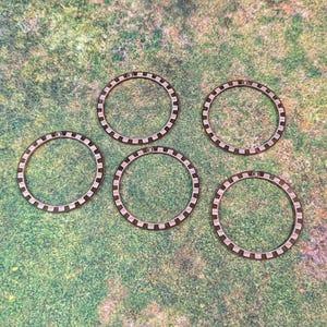 Peut inclure: Cinq jetons circulaires en bois avec une bordure marron foncé et un motif à carreaux au centre. Chaque jeton porte une petite marque circulaire. Les jetons sont disposés sur une surface verte texturée.