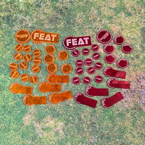 May include: A collection of orange and red game tokens and markers. The tokens feature circular and rectangular shapes with the words "FEAT", "FOCUS", "FURY", and "SPELL". They are arranged on a green surface.