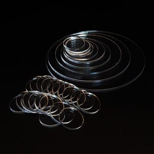 May include: Clear acrylic circles and rings of varying sizes are arranged against a black background. The circles are stacked in a spiral pattern, while the rings are scattered in a separate pile. The transparent material reflects light, creating highlights and shadows.
