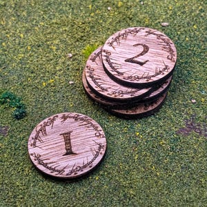 May include: Wooden tokens with the numbers 1 and 2 engraved on them. The tokens are circular and have a decorative border. The tokens are on a green surface.