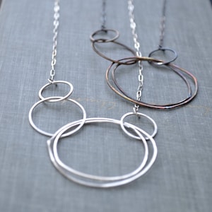 May include: A silver necklace with multiple interconnected circles, featuring a larger circle with a smaller circle inside it. The necklace is on a gray textured surface.