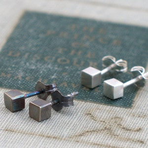 May include: Two pairs of earrings, one pair is silver and the other is a dark grey metal. Both pairs are square shaped.