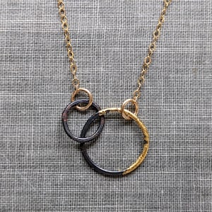 Puede incluir: Un collar de cadena dorada con tres colgantes circulares conectados. Dos de los colgantes son negros con un borde dorado, y el tercero es dorado. El collar se muestra sobre un fondo gris texturizado.