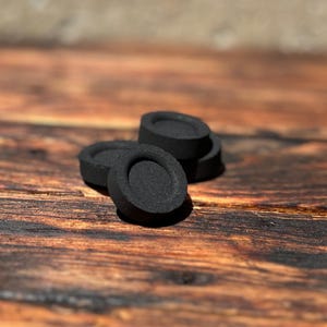 Puede incluir: Varios briquetas de carbón vegetal negras y circulares están dispuestas sobre una superficie de madera. Cada briqueta tiene una hendidura central. Las briquetas son de color negro mate y parecen estar hechas de carbón vegetal comprimido.