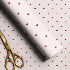 May include: A roll of white wrapping paper featuring a repeating pattern of small red hearts, accompanied by a pair of gold-toned metal scissors with ornate handle detailing, positioned diagonally across the paper surface.